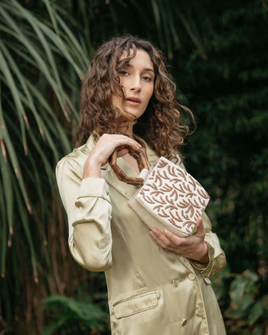 Model in a cream jumpsuit holding the Symbolic Path beaded bag in a lush green garden setting, showcasing its elegant handheld style.