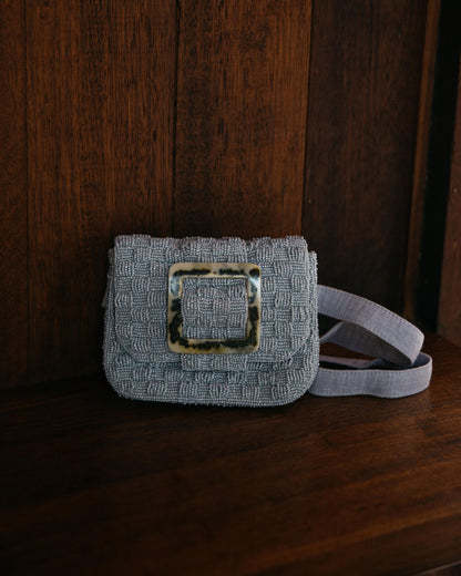 A lifestyle mood shot of the soft blue Uncovered Layers waist bag, showcasing its artisanal hand-dyed glass beads in a natural setting.